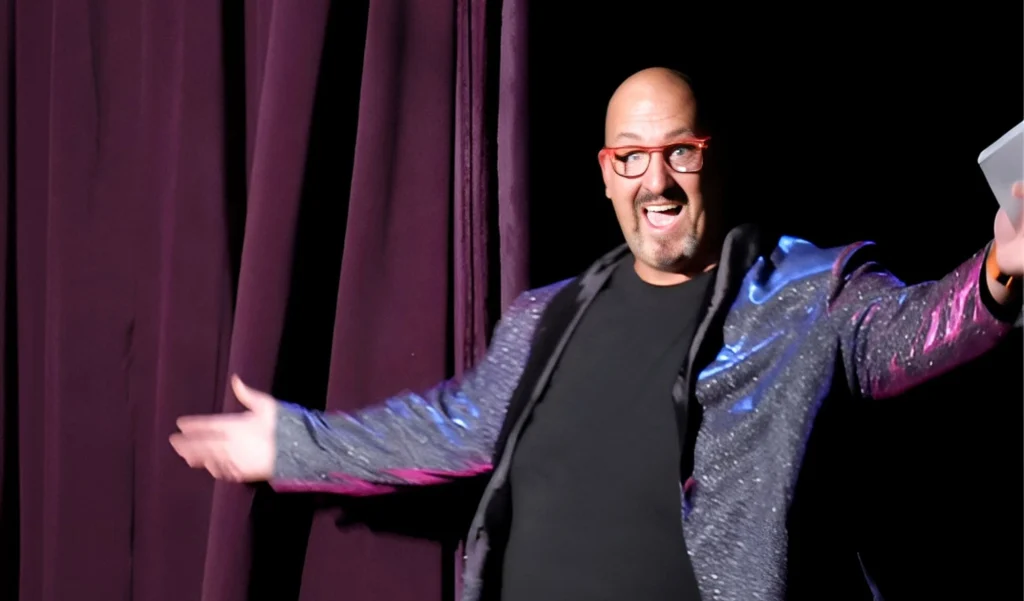 Jon Evans on a dark stage in a sparkly purple jacket and red glasses, arms spread wide with a big smile.