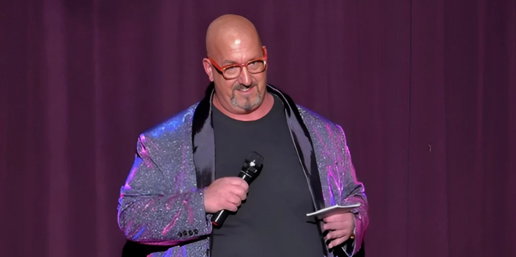 Jon Evans on stage in a sparkly jacket and red glasses, holding a microphone and a note card.
