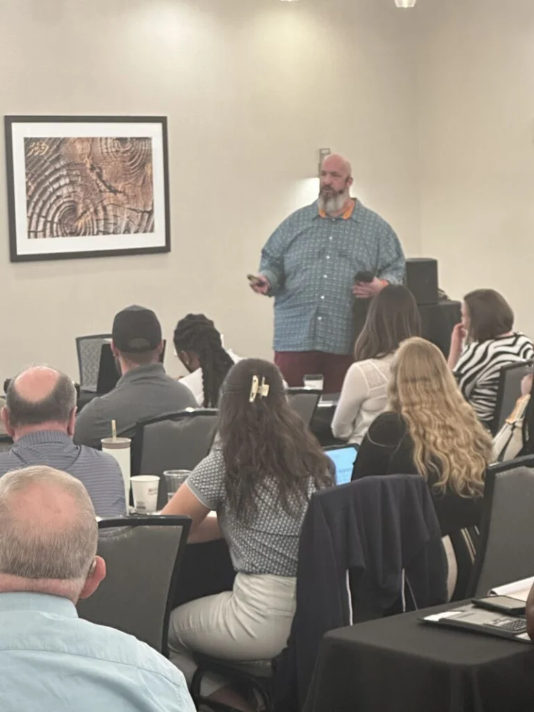 Jon Evans standing at the front of a meeting room, presenting to attendees seated at tables.