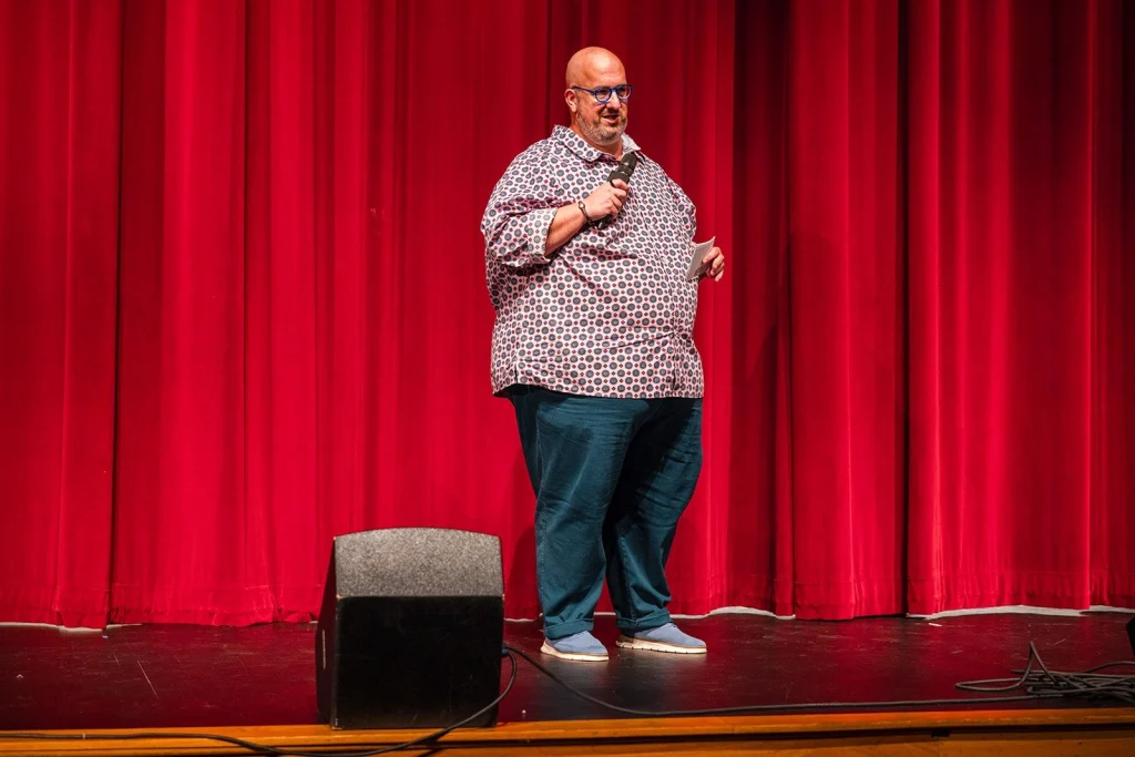 Jon Evans standing on stage in front of a red curtain, holding a microphone and a note card while emceeing an event.